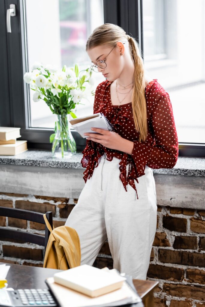 attractive student in glasses writing in notebook in apartment